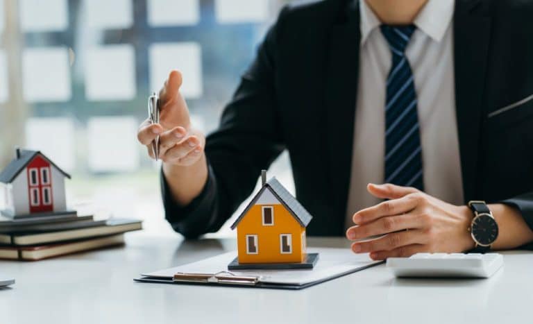 Une personne en costume discute d'investissement immobilier à un bureau avec une maison modèle, un presse papiers et une calculatrice.