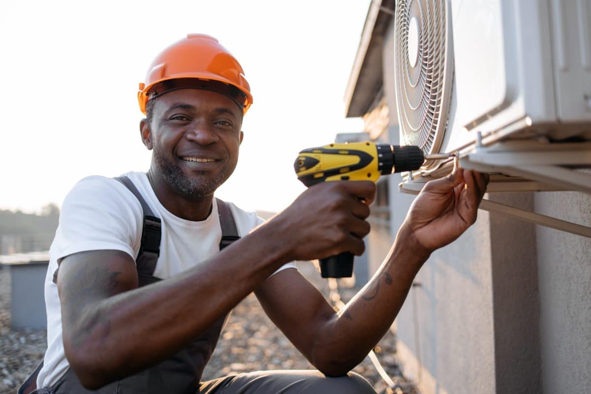 Une personne portant un casque de sécurité utilise une perceuse sans fil pour travailler sur une unité de climatisation extérieure. Man using screwdriver for repairing ventilation system