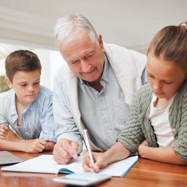 Un homme âgé aide deux enfants à faire leurs devoirs à une table, avec un ordinateur portable et un cahier devant eux.