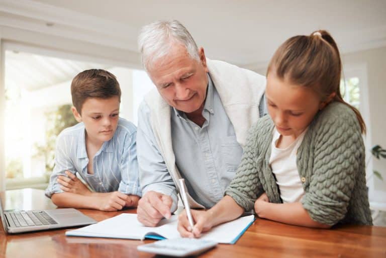 Un homme âgé aide deux enfants à faire leurs devoirs à une table, avec un ordinateur portable et un cahier devant eux.