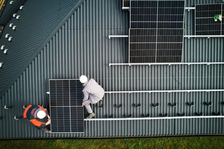 Deux ouvriers casqués installent des panneaux solaires sur un toit métallique incliné.