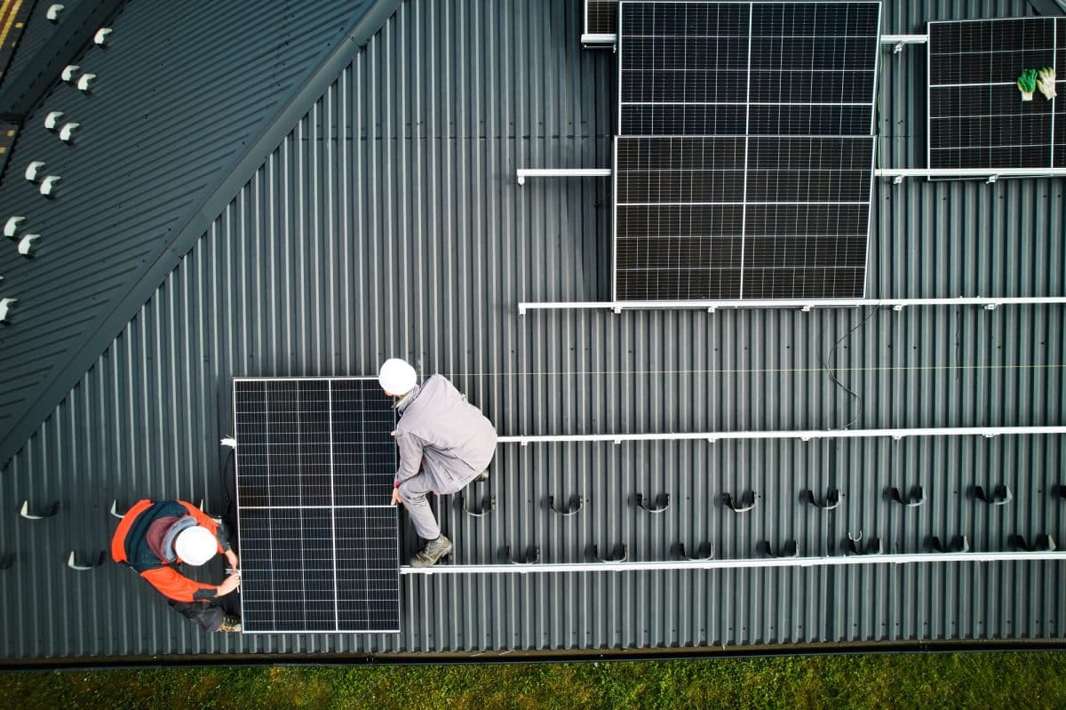 Deux ouvriers casqués installent des panneaux solaires sur un toit métallique incliné.