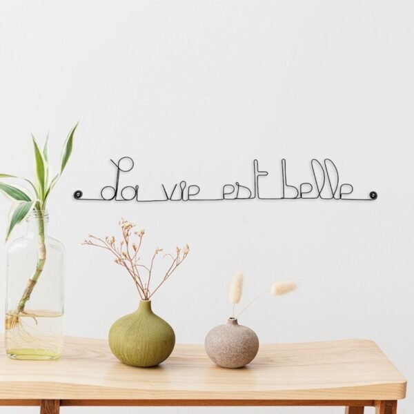 Scène de décoration minimaliste avec un panneau en fil de fer "La vie est belle" sur un mur blanc au dessus d'une table en bois contenant trois vases avec des plantes et des fleurs séchées.