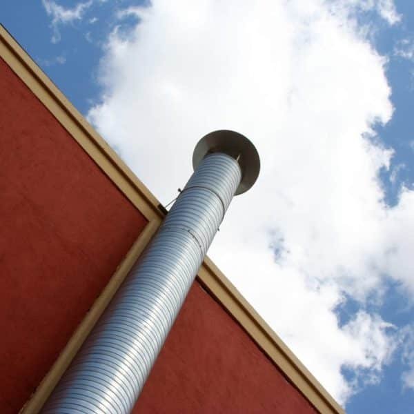 Vue d'en bas d'une haute cheminée en métal argenté sur un bâtiment rouge, avec un ciel bleu nuageux en arrière plan.