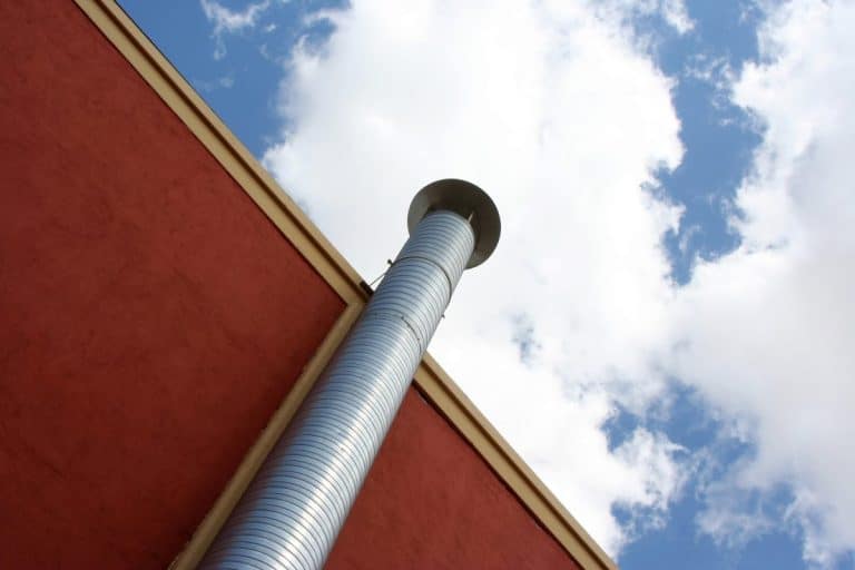 Vue d'en bas d'une haute cheminée en métal argenté sur un bâtiment rouge, avec un ciel bleu nuageux en arrière plan.