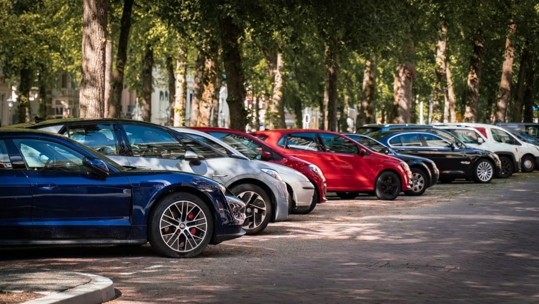Des voitures de différentes couleurs et de différents modèles sont garées en rangée le long d'une rue bordée d'arbres par une journée ensoleillée.
