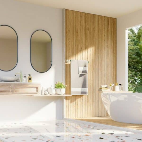 Salle de bain moderne avec baignoire autoportante, deux miroirs ovales et une vasque murale. Une grande fenêtre offre une vue sur le jardin luxuriant. Le bois clair et les tons beiges dominent le décor.