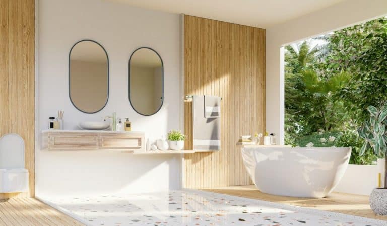 Salle de bain moderne avec baignoire autoportante, deux miroirs ovales et une vasque murale. Une grande fenêtre offre une vue sur le jardin luxuriant. Le bois clair et les tons beiges dominent le décor.