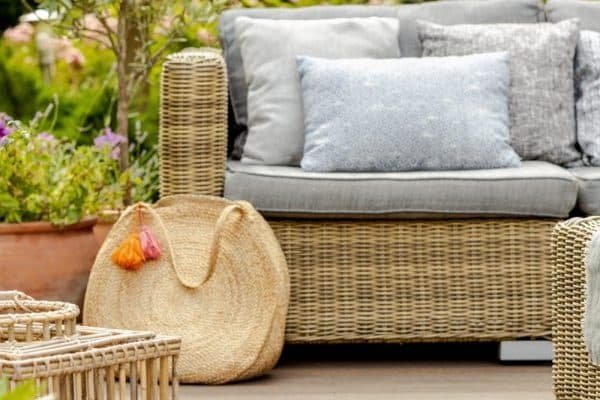 Mobilier de jardin en osier avec coussins gris, un panier tressé, un plateau avec des raisins et une bougie et des plantes en pot dans un décor de jardin.