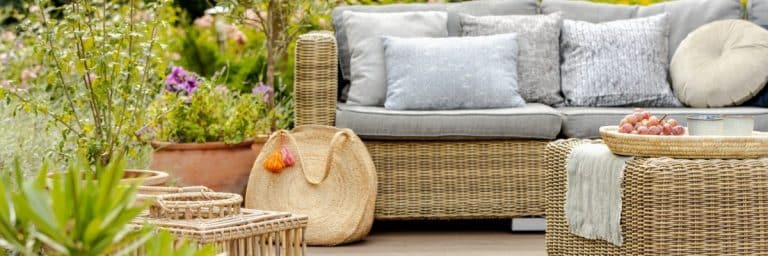 Mobilier de jardin en osier avec coussins gris, un panier tressé, un plateau avec des raisins et une bougie et des plantes en pot dans un décor de jardin.