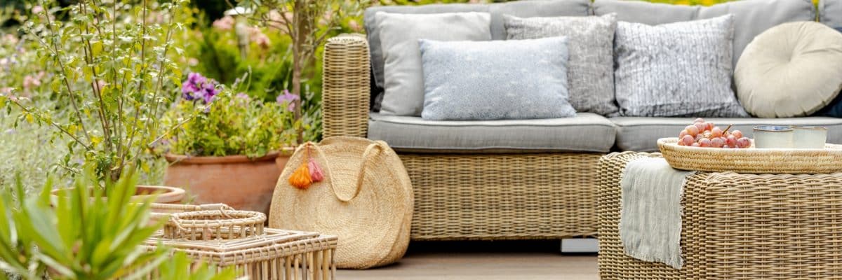 Mobilier de jardin en osier avec coussins gris, un panier tressé, un plateau avec des raisins et une bougie et des plantes en pot dans un décor de jardin.