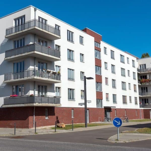 Immeuble d'appartements moderne en briques blanches et rouges avec plusieurs balcons, situé au coin d'une rue, sous un ciel bleu clair.