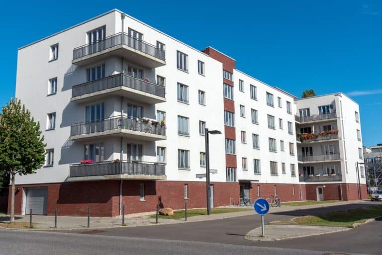 Immeuble d'appartements moderne en briques blanches et rouges avec plusieurs balcons, situé au coin d'une rue, sous un ciel bleu clair.