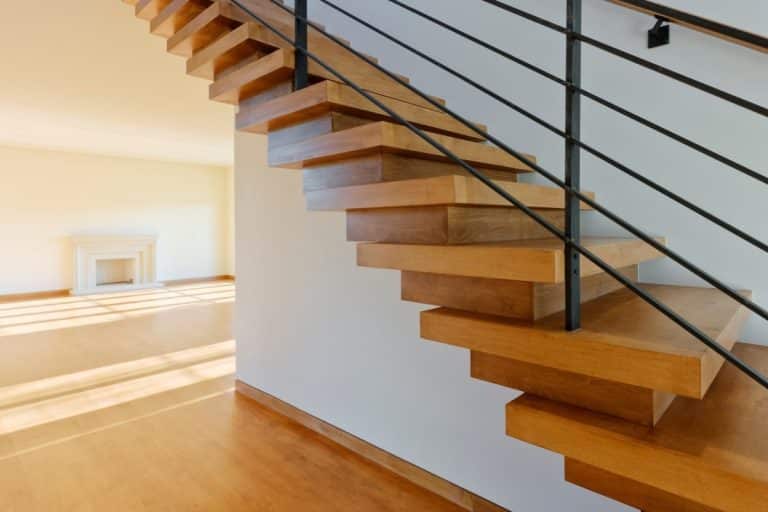 Escalier flottant en bois avec rampe en métal noir, menant à un étage supérieur. La lumière du soleil filtre dans la pièce, mettant en valeur les lignes épurées et le design minimaliste. Une cheminée est visible à proximité.