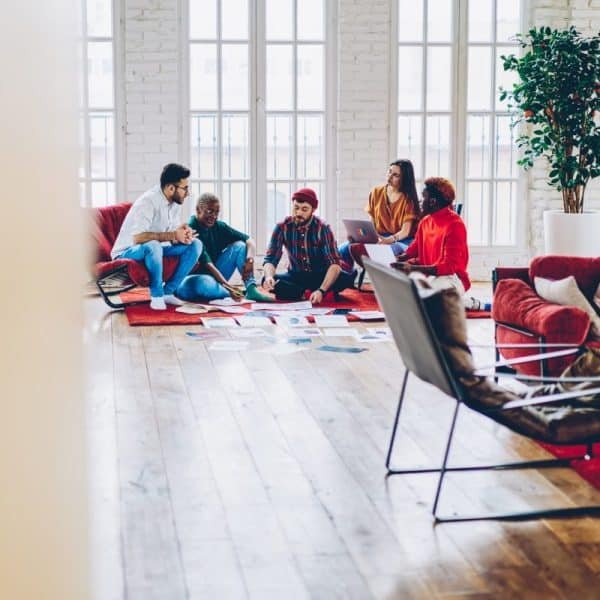 Cinq personnes sont assises par terre et sur un canapé dans un salon très éclairé, discutant de papiers étalés sur le sol et utilisant un ordinateur portable.
