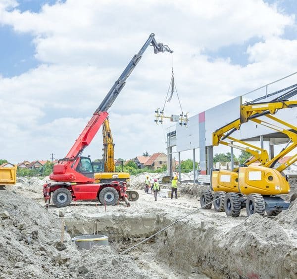 Chantier de construction avec des grues et des machines lourdes soulevant des matériaux près d'une structure partiellement construite, tandis que des ouvriers se tiennent à proximité sous un ciel partiellement nuageux.