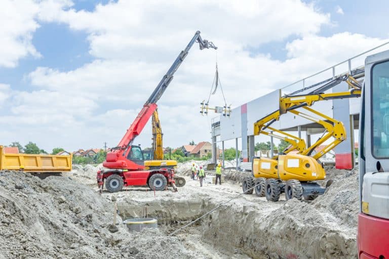 Chantier de construction avec des grues et des machines lourdes soulevant des matériaux près d'une structure partiellement construite, tandis que des ouvriers se tiennent à proximité sous un ciel partiellement nuageux.