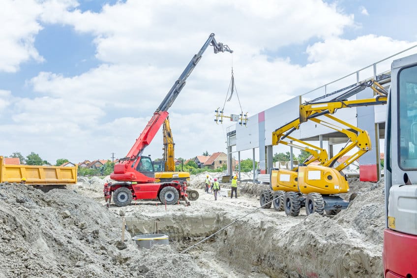 Chantier de construction avec des grues et des machines lourdes soulevant des matériaux près d'une structure partiellement construite, tandis que des ouvriers se tiennent à proximité sous un ciel partiellement nuageux.