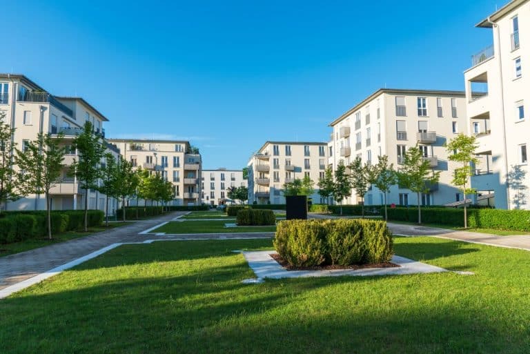 Des immeubles d'appartements modernes entourent un espace vert central avec des sentiers et des buissons sous un ciel bleu clair.