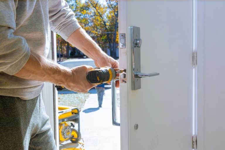 Une personne utilisant une perceuse électrique pour installer ou réparer une serrure de porte sur une porte blanche tandis que la lumière du soleil brille à travers la porte ouverte.