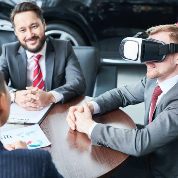 Trois hommes en costume sont assis autour d'une table. L'un d'eux porte un casque de réalité virtuelle, tandis qu'un autre sourit et que le troisième consulte un document. Une voiture et des fleurs sont visibles en arrière plan.