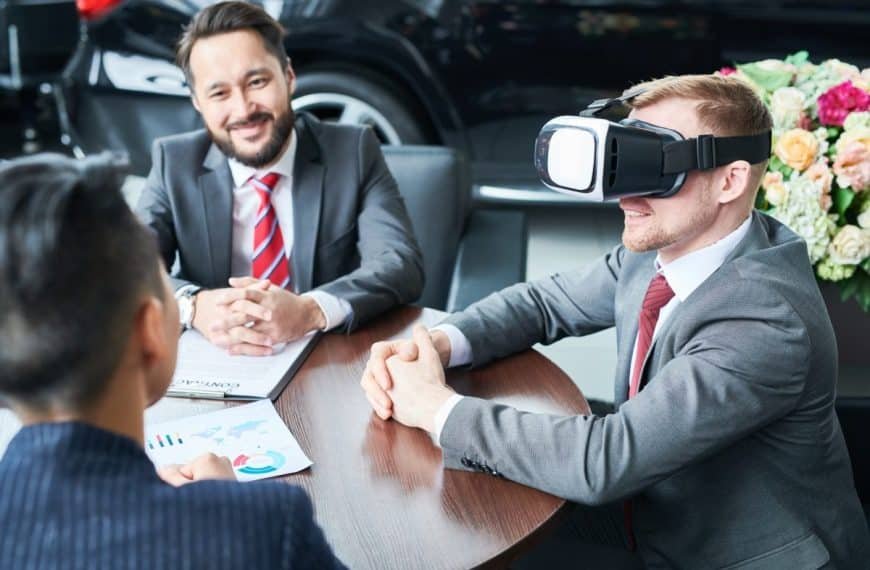 Trois hommes en costume sont assis autour d'une table. L'un d'eux porte un casque de réalité virtuelle, tandis qu'un autre sourit et que le troisième consulte un document. Une voiture et des fleurs sont visibles en arrière plan.