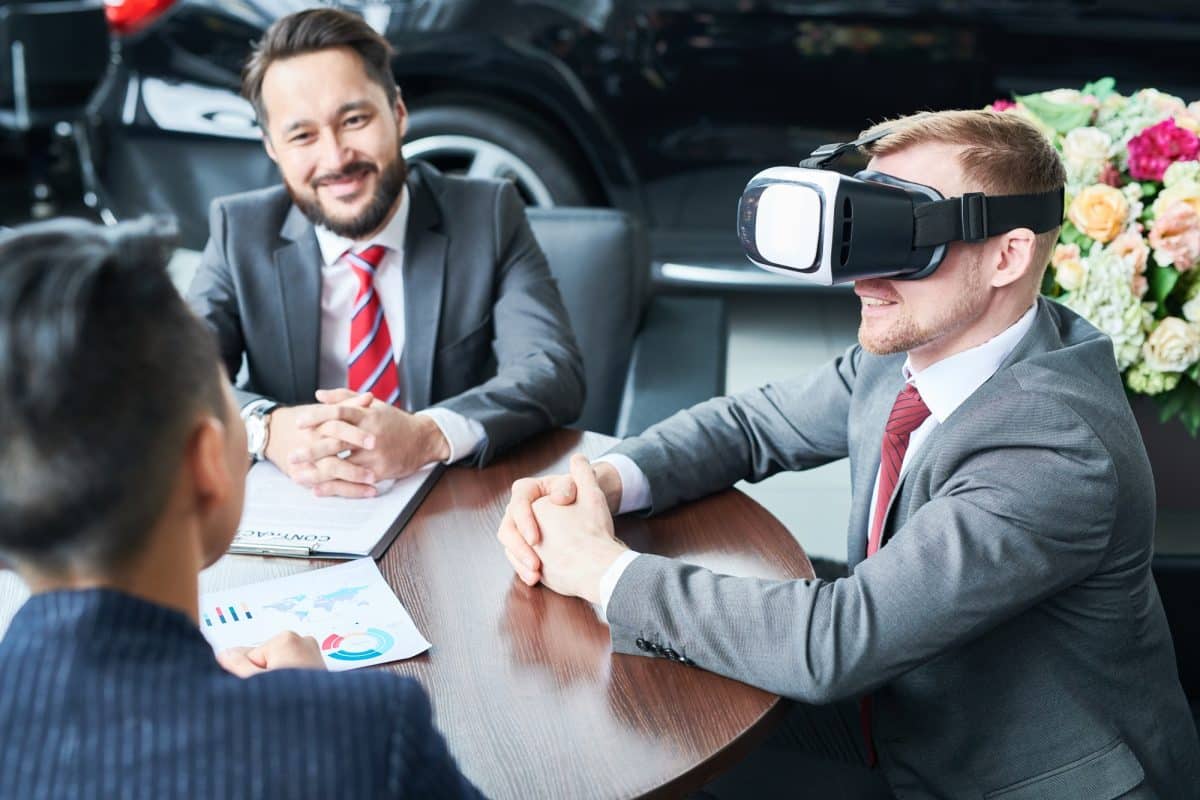 Trois hommes en costume sont assis autour d'une table. L'un d'eux porte un casque de réalité virtuelle, tandis qu'un autre sourit et que le troisième consulte un document. Une voiture et des fleurs sont visibles en arrière plan.