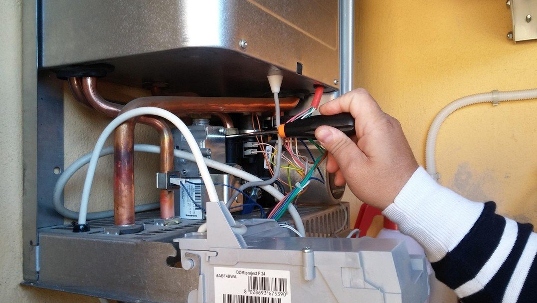 Une personne utilise un tournevis pour travailler sur le câblage électrique à l'intérieur d'un appareil métallique ouvert monté sur un mur.
