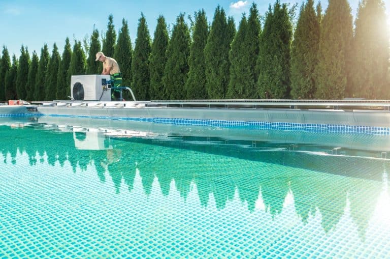 Une personne portant un chapeau travaille sur un système de pompe de piscine à côté d'une grande piscine extérieure, avec une rangée de grands arbres verts en arrière plan sous un ciel bleu.