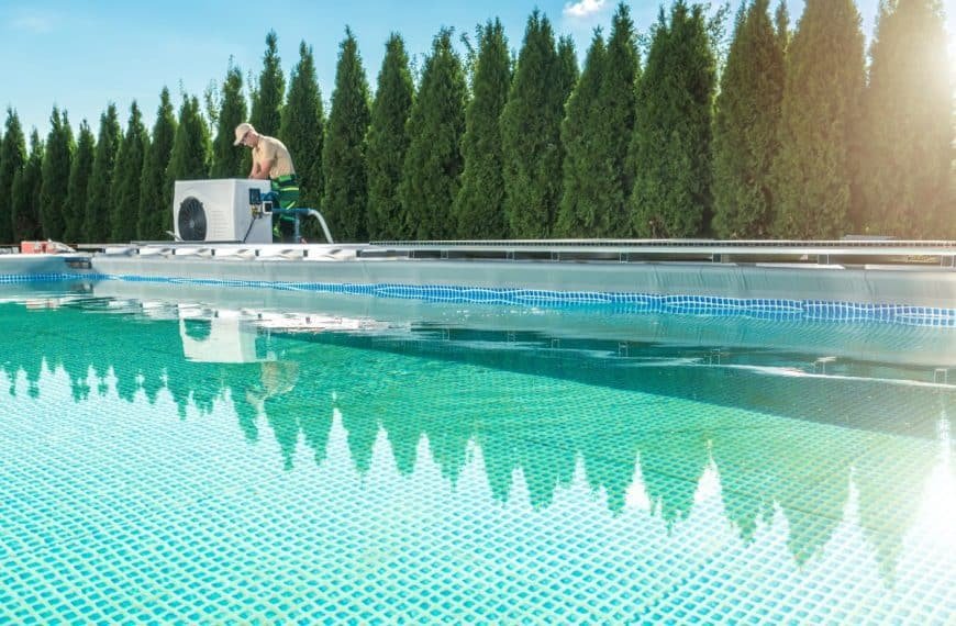 Une personne portant un chapeau travaille sur un système de pompe de piscine à côté d'une grande piscine extérieure, avec une rangée de grands arbres verts en arrière plan sous un ciel bleu.