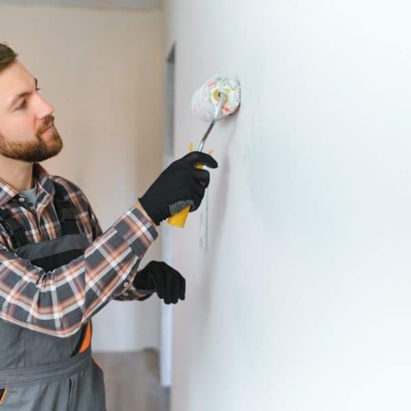 Un homme portant une chemise à carreaux et une salopette, utilisant un rouleau à peinture pour peindre un mur blanc.