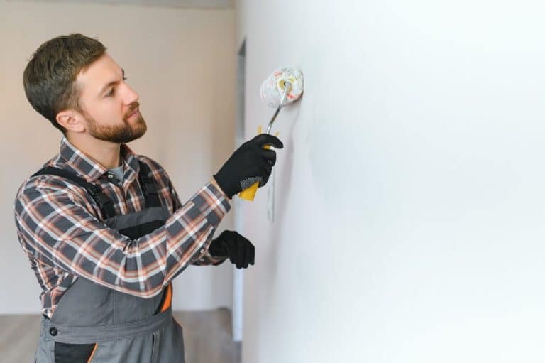 Un homme portant une chemise à carreaux et une salopette, utilisant un rouleau à peinture pour peindre un mur blanc.