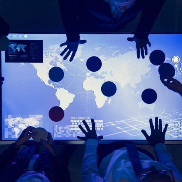 Les gens se tiennent autour d'une table numérique affichant une carte du monde, plaçant des disques colorés à divers endroits, suggérant une séance de planification collaborative ou stratégique.
