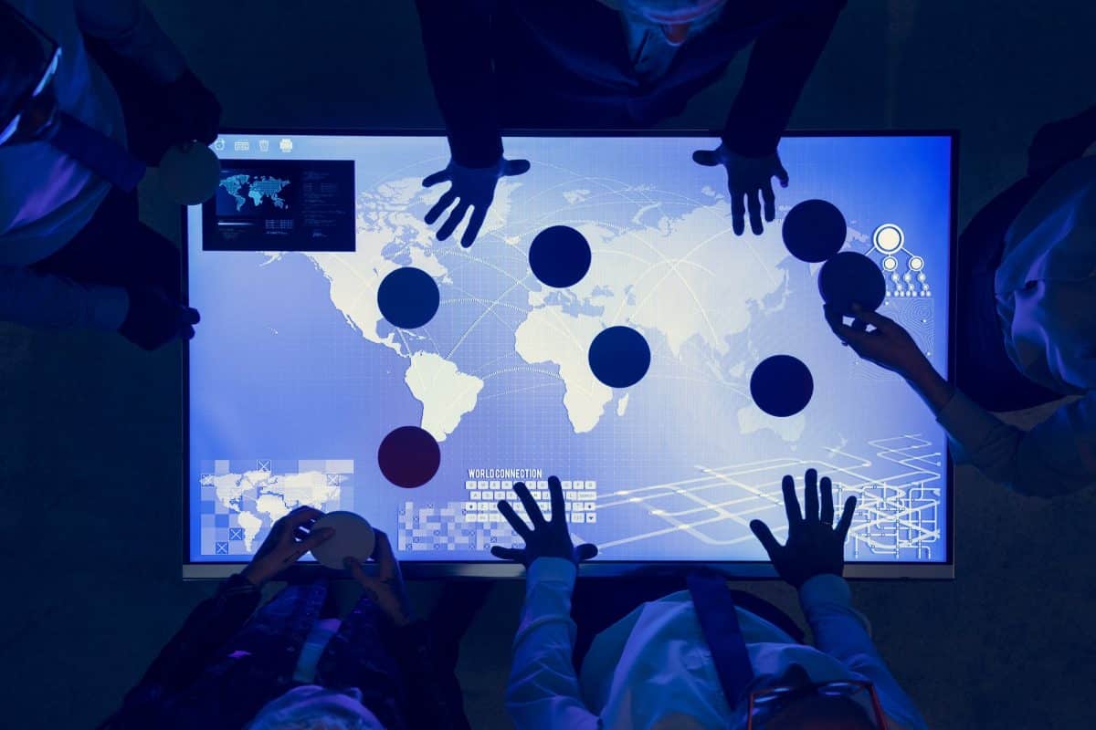 Les gens se tiennent autour d'une table numérique affichant une carte du monde, plaçant des disques colorés à divers endroits, suggérant une séance de planification collaborative ou stratégique.