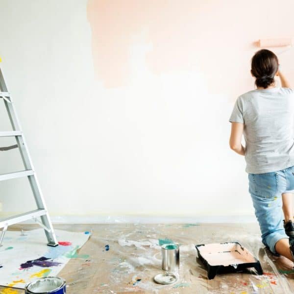 Une personne peint un mur couleur pêche, agenouillée au sol. On aperçoit une échelle, un bac à peinture et une bâche recouvrant le sol.