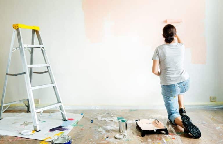 Une personne peint un mur couleur pêche, agenouillée au sol. On aperçoit une échelle, un bac à peinture et une bâche recouvrant le sol.