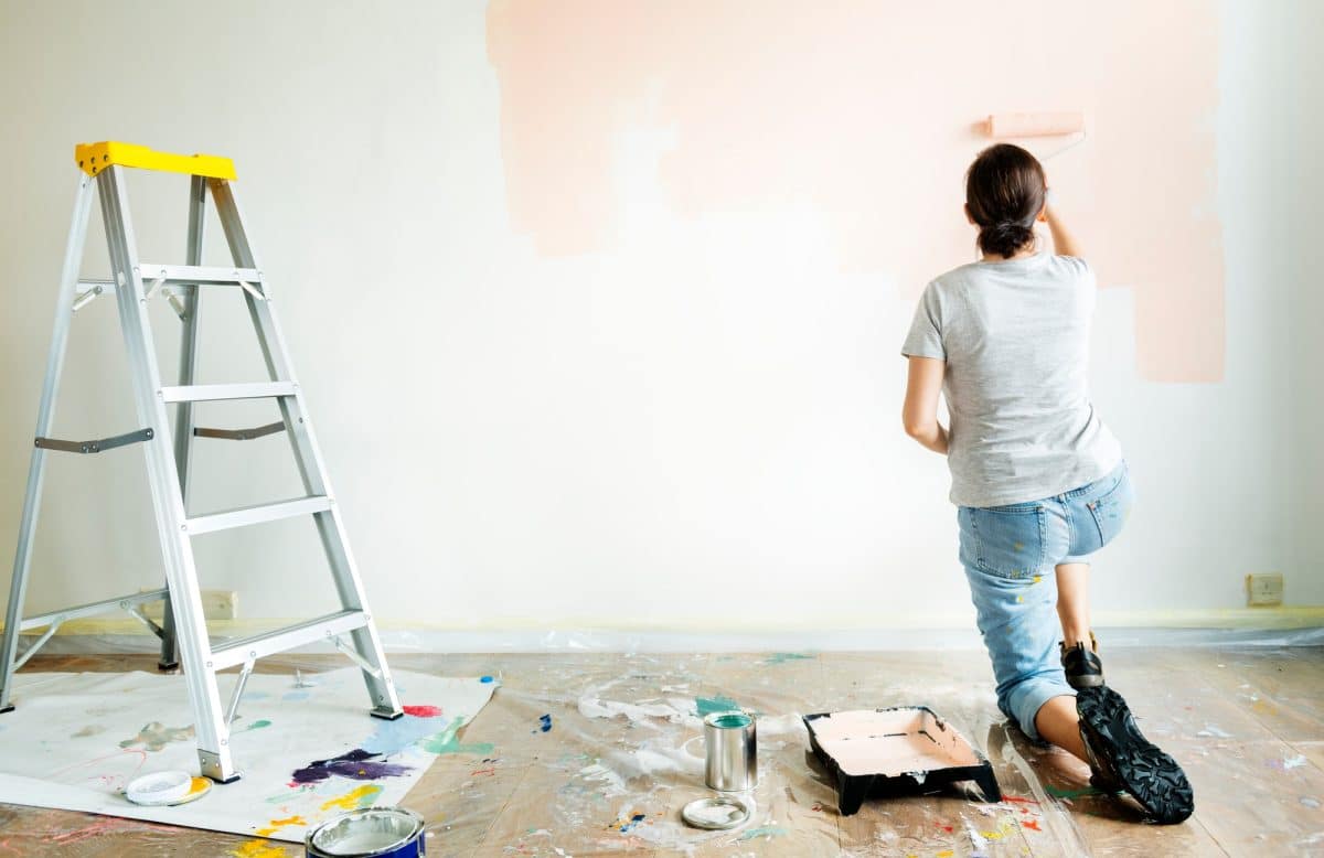 Une personne peint un mur couleur pêche, agenouillée au sol. On aperçoit une échelle, un bac à peinture et une bâche recouvrant le sol.