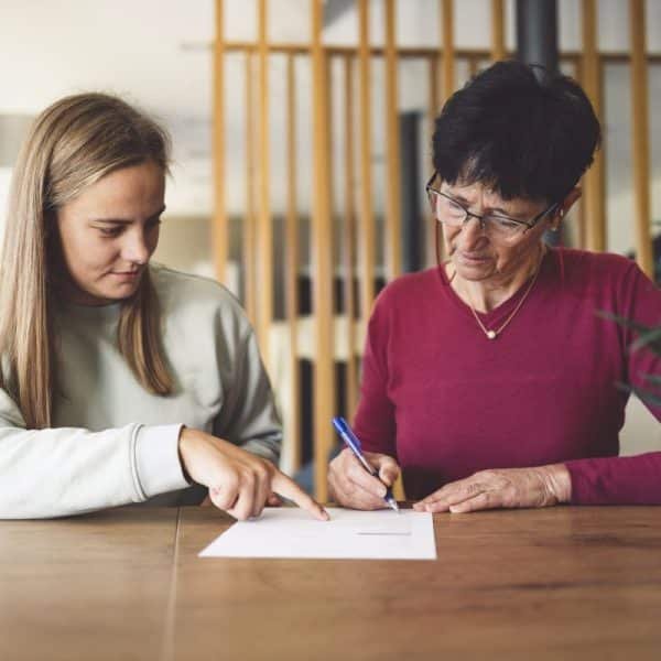 Deux femmes sont assises à une table ; l'une signe un document tandis que l'autre montre du doigt le papier, toutes deux concentrées sur leur tâche.
