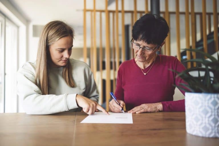 Deux femmes sont assises à une table ; l'une signe un document tandis que l'autre montre du doigt le papier, toutes deux concentrées sur leur tâche.