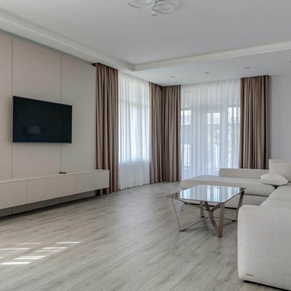 Salon moderne avec télévision murale, canapé d'angle beige, table basse en verre, lampadaire et grandes fenêtres avec rideaux marron et blancs. Un parquet en bois clair complète le design minimaliste.