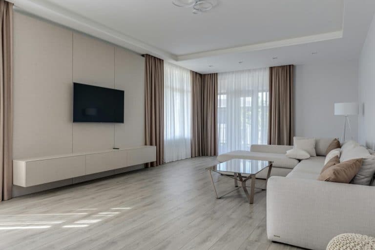 Salon moderne avec télévision murale, canapé d'angle beige, table basse en verre, lampadaire et grandes fenêtres avec rideaux marron et blancs. Un parquet en bois clair complète le design minimaliste.
