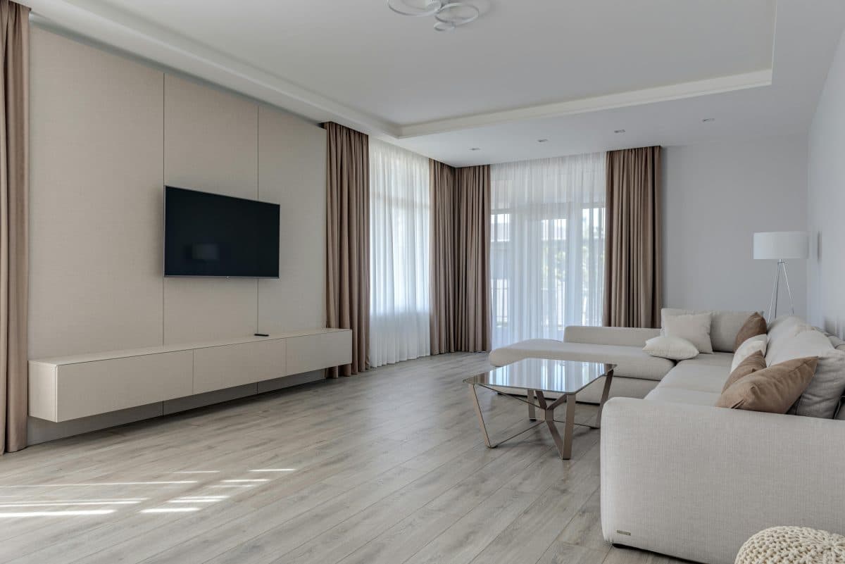 Salon moderne avec télévision murale, canapé d'angle beige, table basse en verre, lampadaire et grandes fenêtres avec rideaux marron et blancs. Un parquet en bois clair complète le design minimaliste.