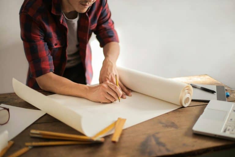 Une personne portant une chemise à carreaux rouge écrit sur une grande feuille de papier posée sur une table en bois, entourée de crayons et de plans, avec un ordinateur portable ouvert à proximité.