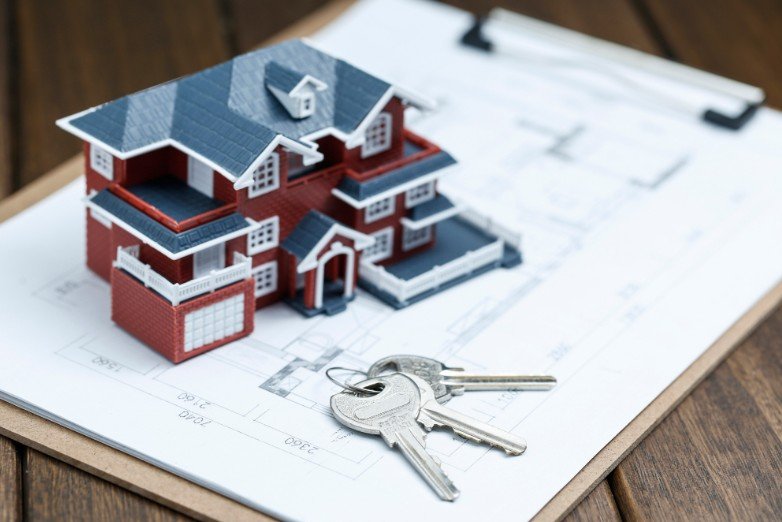 Une maquette de maison et un trousseau de clés reposent sur des plans d'architecture accrochés à un presse-papiers en bois sur une surface en bois.