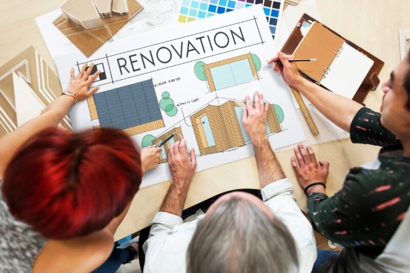Trois personnes discutent d'un plan de rénovation à une table où se trouvent des échantillons de couleurs, un carnet de notes et des maquettes de bâtiments.