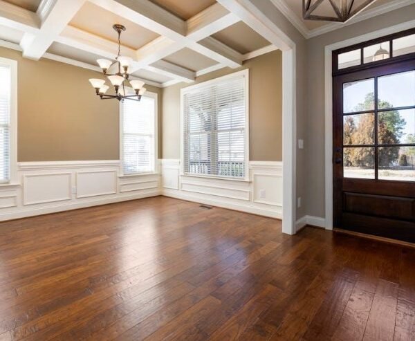 Pièce vide avec un sol en bois, des murs beiges, des boiseries blanches, de grandes fenêtres avec des stores, un lustre et une porte vitrée donnant sur l'extérieur.