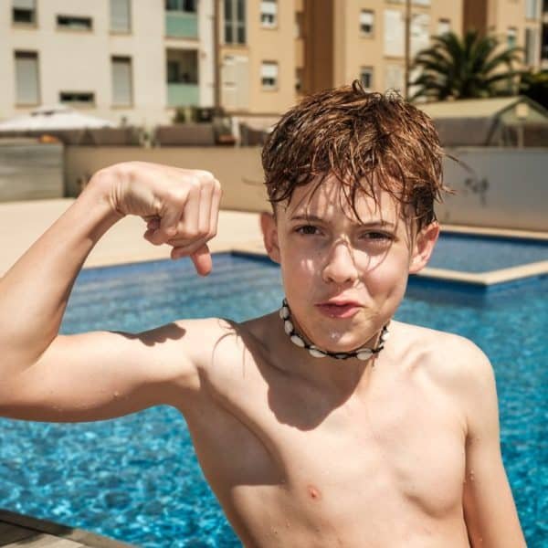 Un garçon se tient au bord de la piscine, les cheveux mouillés et torse nu, fléchissant le bras et affichant une grimace enjouée. Des immeubles d'habitation sont visibles en arrière plan.