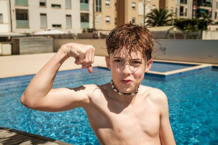 Un garçon se tient au bord de la piscine, les cheveux mouillés et torse nu, fléchissant le bras et affichant une grimace enjouée. Des immeubles d'habitation sont visibles en arrière plan.