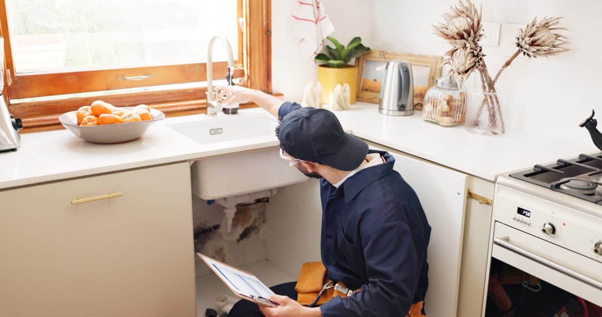 Un homme portant une casquette et une salopette examine le robinet d'un évier de cuisine, un presse papiers à la main. Il y a un bol d'oranges sur le comptoir à côté de lui. Plumber, house and handyman with clipboard, maintenance and service with expert. Employee, contract