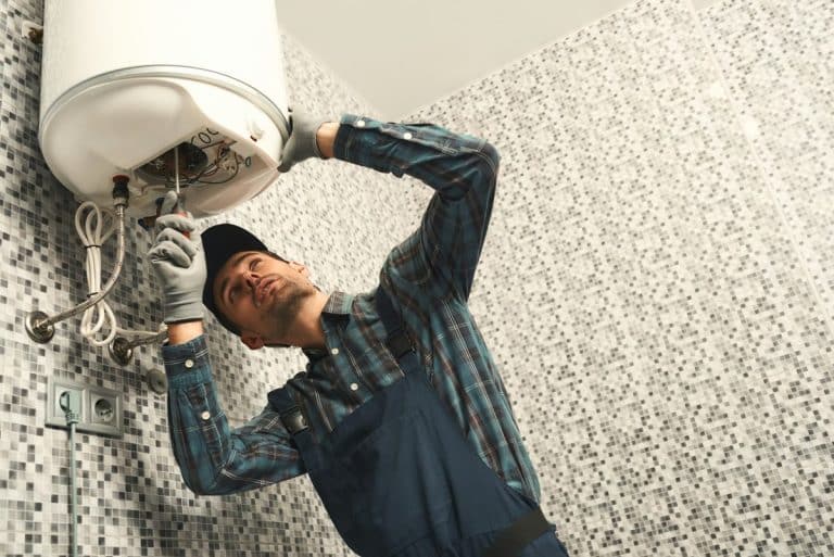 Un homme portant une chemise à carreaux, une salopette et une casquette est en train de réparer un chauffe-eau mural dans une pièce carrelée.